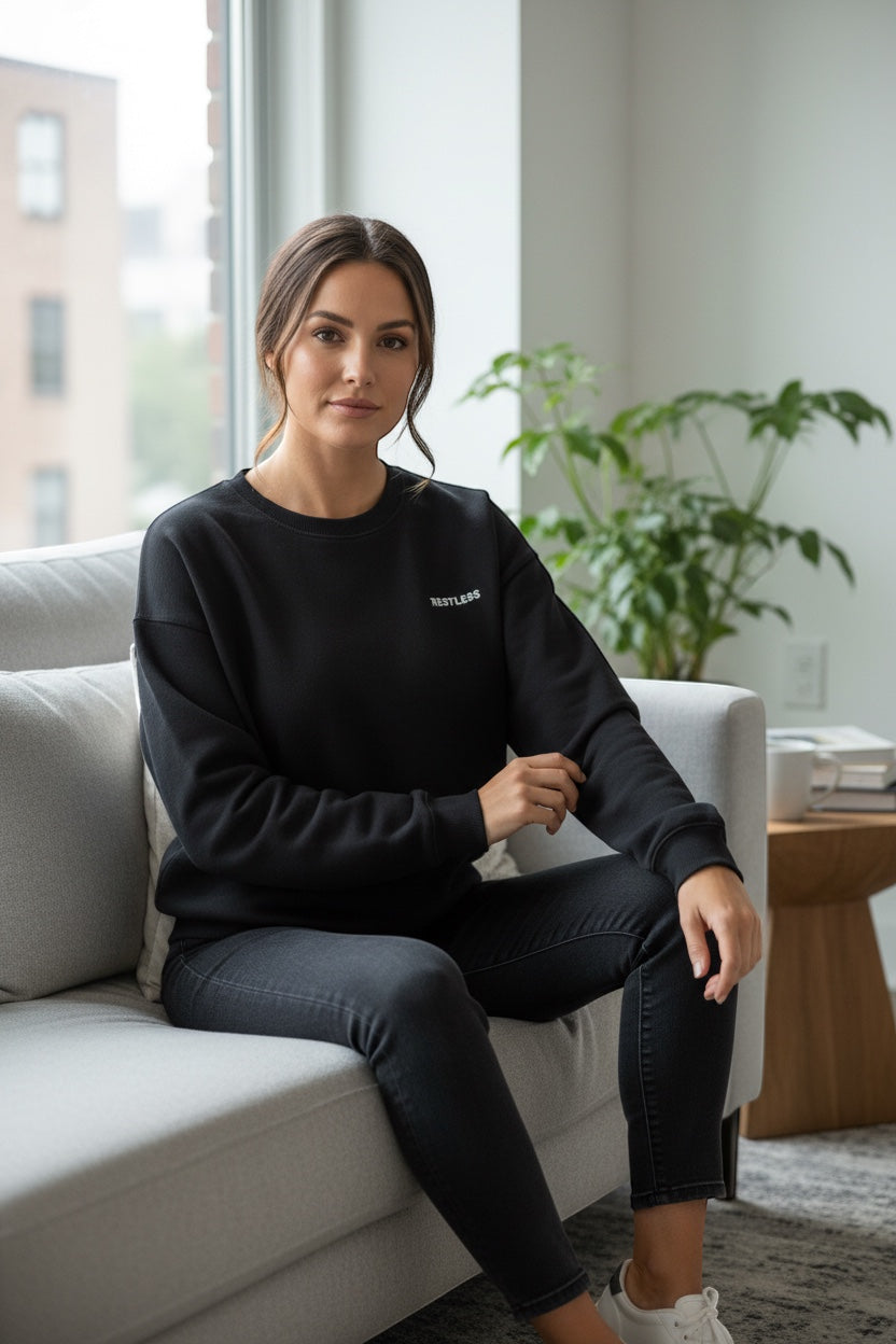 Restless black crewneck sweatshirt made of flannel fabric, featuring a small embroidered logo on the chest. Worn by a woman sitting on a couch.