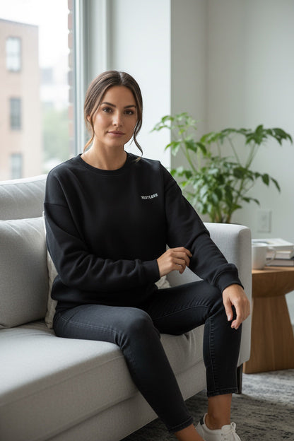 Restless black crewneck sweatshirt made of flannel fabric, featuring a small embroidered logo on the chest. Worn by a woman sitting on a couch.
