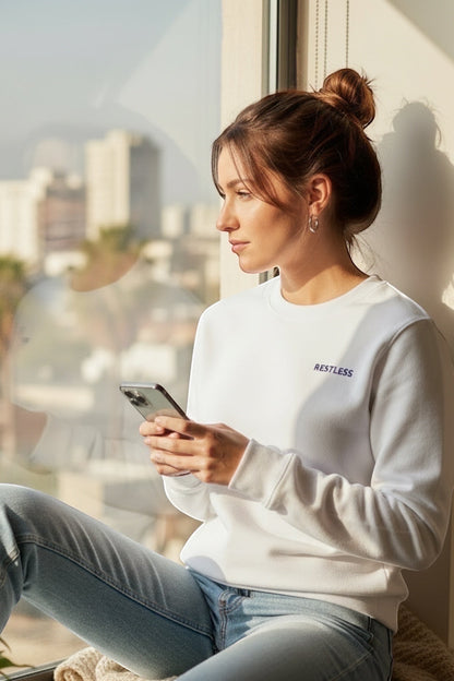 Restless white classic sweatshirt made of flannel fabric with embroidered logo, worn by a woman sitting by a window.